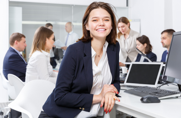 An office setting, people gathered around table, a woman is smiling at the front