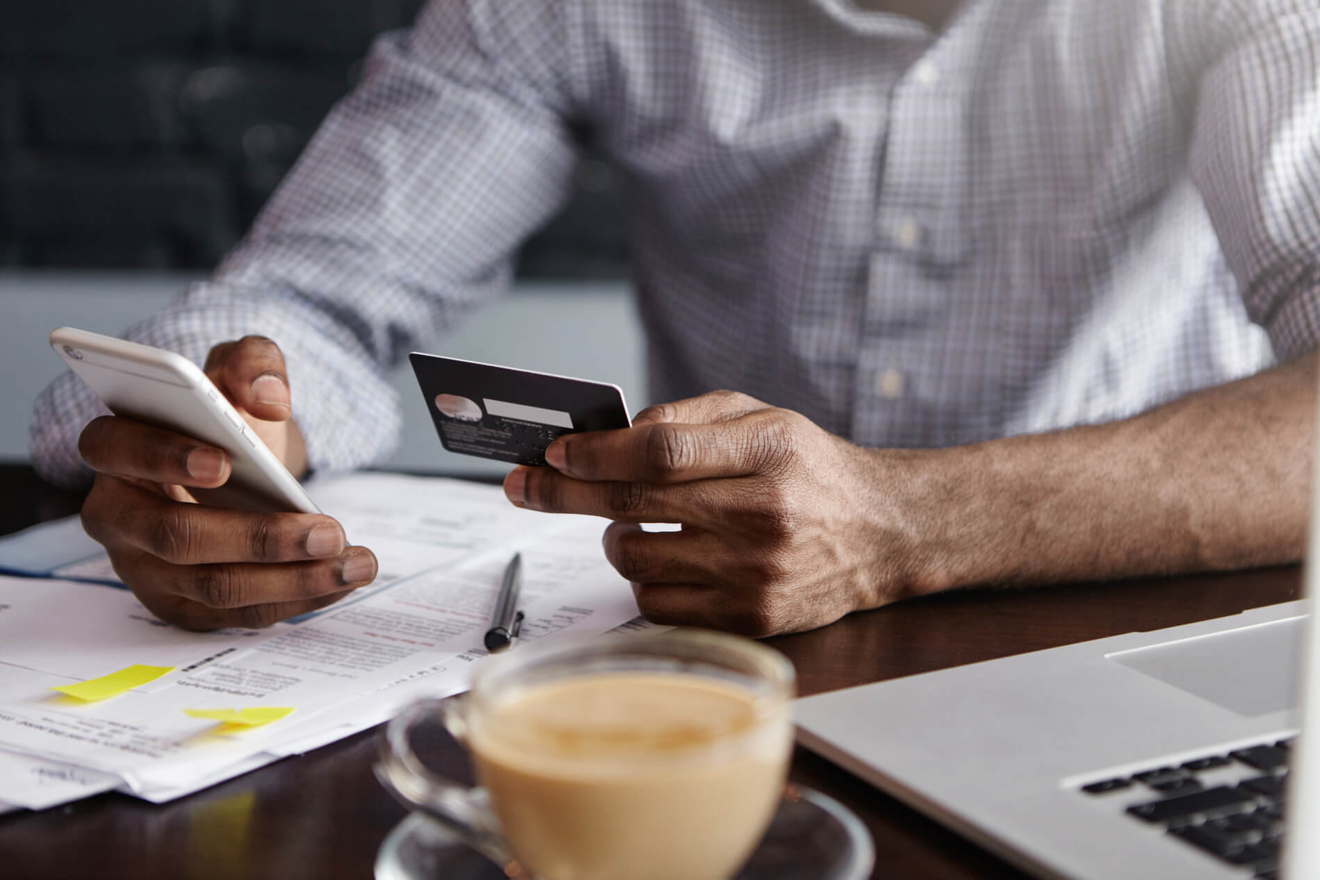 A man holding credit card in one hand and a smartphone in another hand, coffee cup on a desk